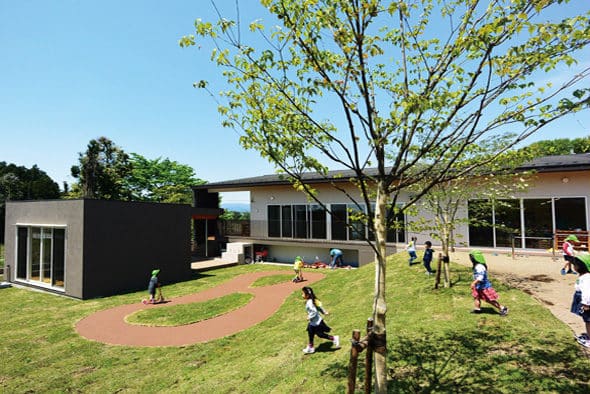 芝生の園庭を走り回る子どもたち。奥に濃茶の木板張りと白い壁を組み合わせた平屋の園舎
