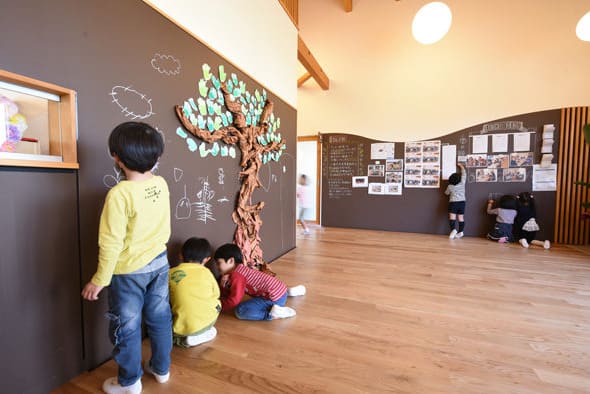 黒板の壁に描かれた木の絵に葉を貼る子どもたち。木の床の広い保育室