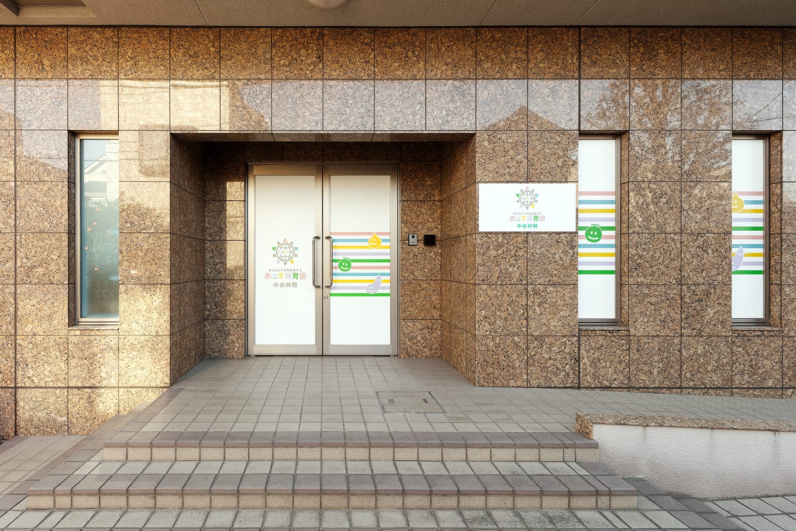 あーす保育園中央林間の玄関の外観写真。園の看板と窓とドアのガラスに看板模様のフィルムを張っている。